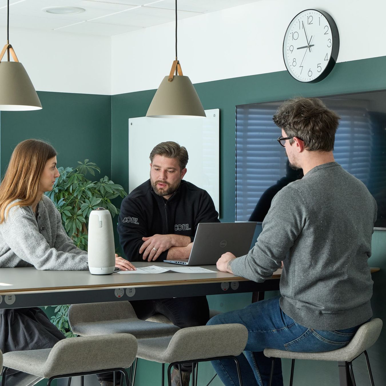 Three people converse around a desk in a modern office, with laptops and notes. The atmosphere is collaborative, under soft pendant lights and a wall clock