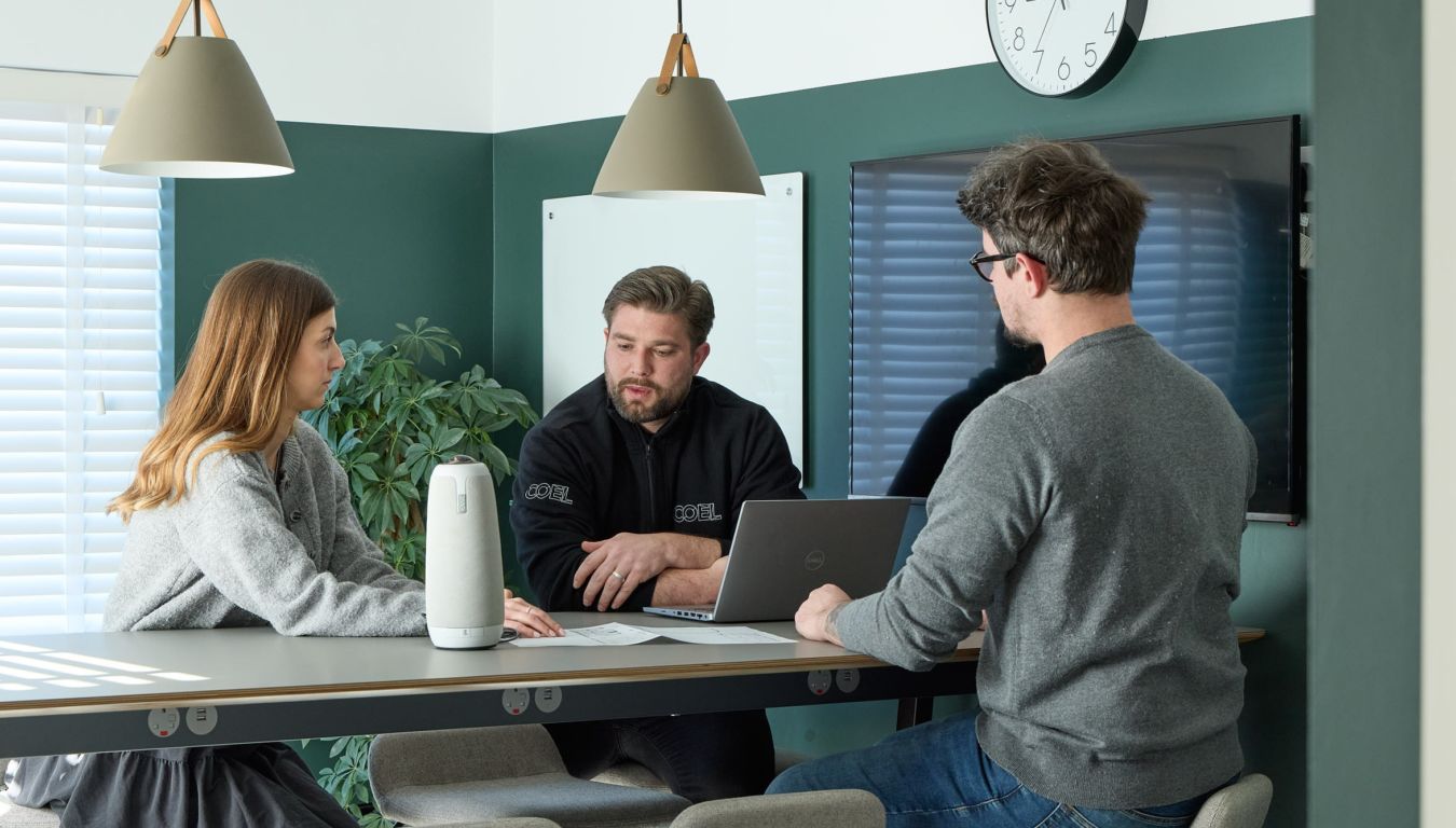 Three people converse around a desk in a modern office, with laptops and notes. The atmosphere is collaborative, under soft pendant lights and a wall clock