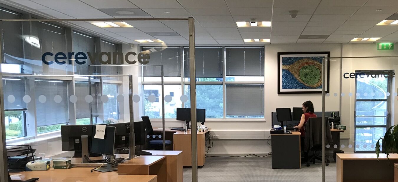 An office environment features desks and computers, with one person working, separated by glass partitions. The text "cerevance" is visible on the glass, and a colorful artwork adorns the wall.