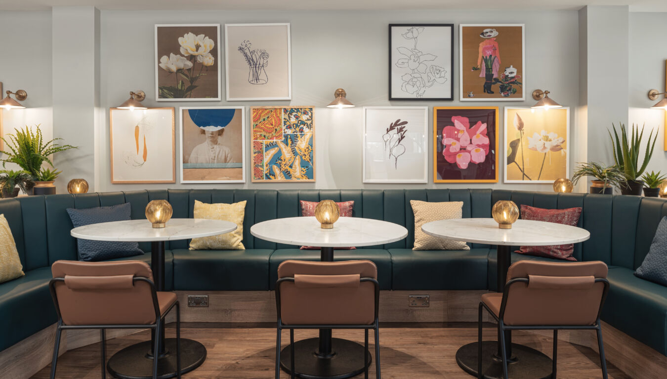 Stylish cafe with three round tables, leather chairs, and teal booth seating. Colorful art on white walls and plants create a cozy, modern atmosphere