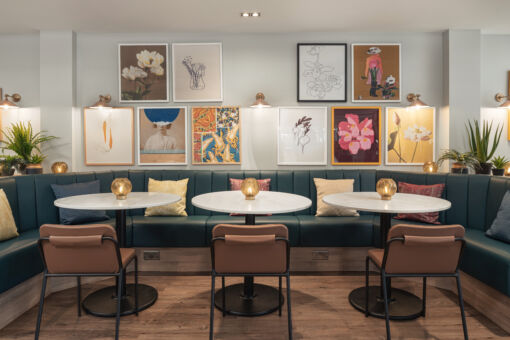 Stylish cafe with three round tables, leather chairs, and teal booth seating. Colorful art on white walls and plants create a cozy, modern atmosphere