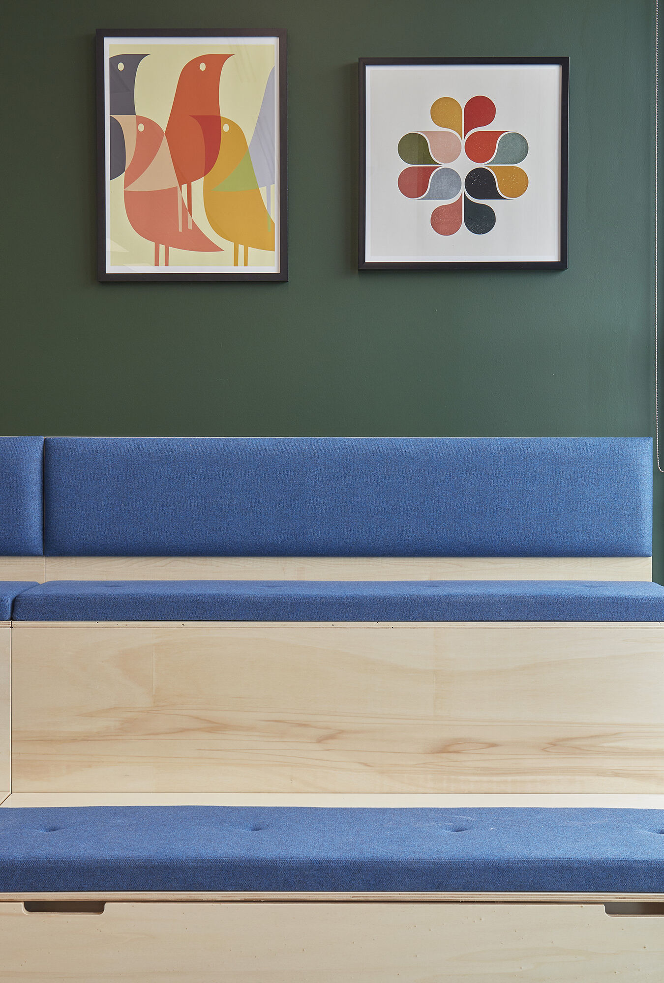 Modular wooden bench with blue cushions against a dark green wall. Two colorful abstract art pieces hang above. Bright, modern, and minimalist