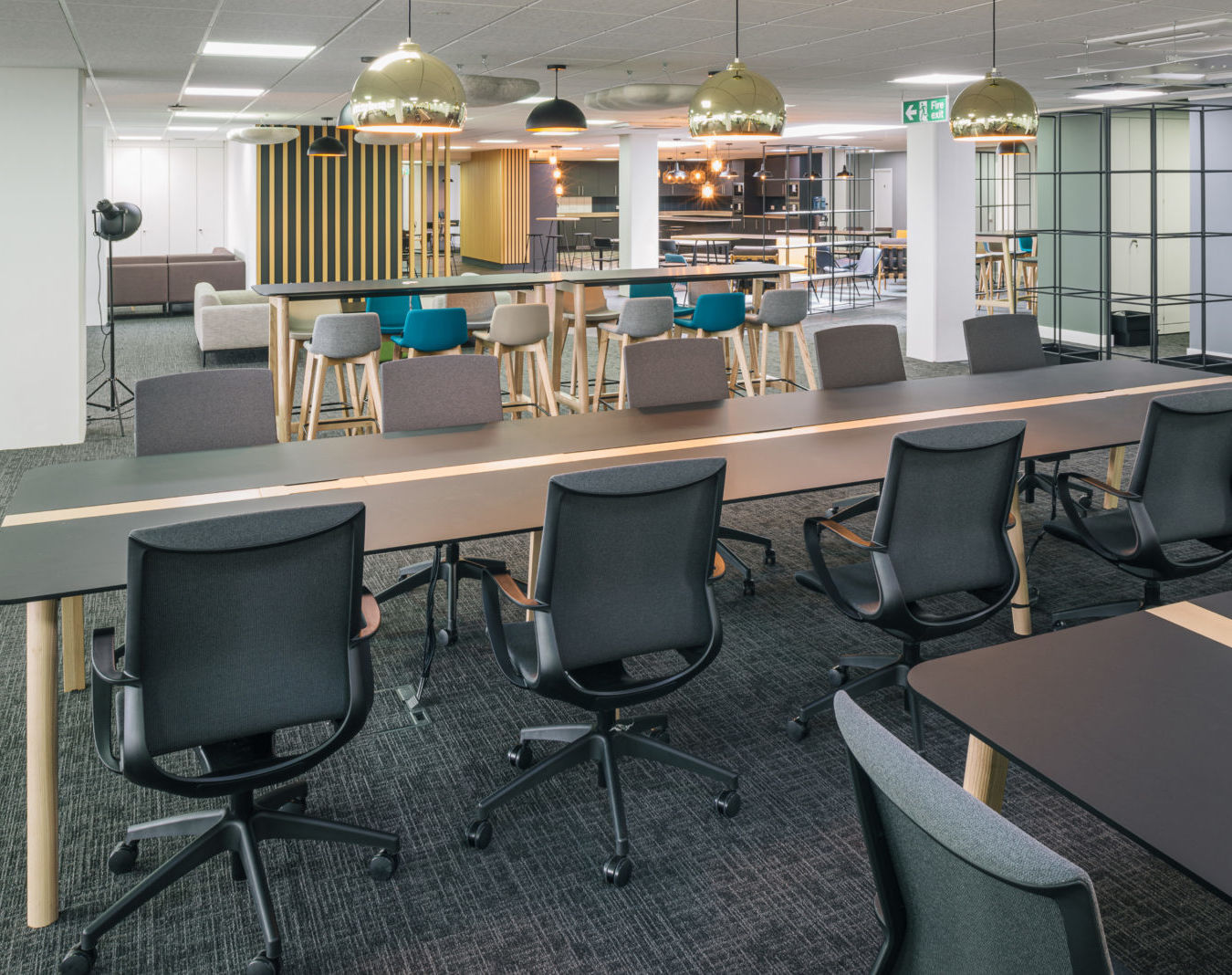 Modern office space with long black tables, ergonomic chairs, and pendant lights. Bar stools and a cozy seating area in the background create a collaborative atmosphere