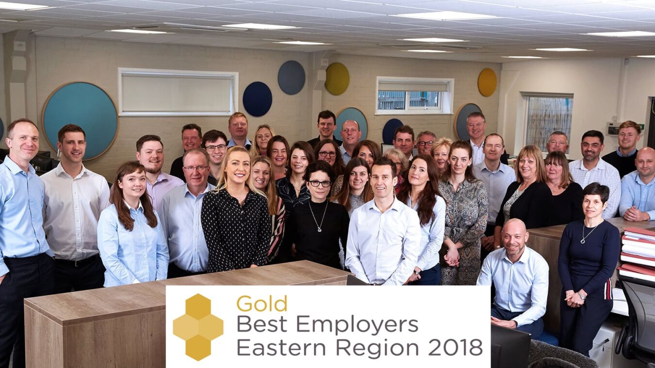 Group of smiling people posing in an office setting with “Gold Best Employers Eastern Region 2018” text overlay at the bottom. Circular wall decor adds a decorative touch.