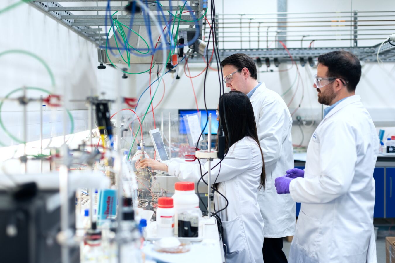 Scientists in lab coats conduct experiments with laboratory equipment in a bright, modern lab. Colored wires hang from the ceiling and various bottles and instruments fill the workspace.
