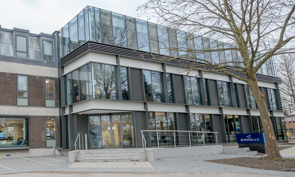 A modern glass-fronted building with steps leading to the entrance, surrounded by trees. A blue sign reads "BIDWELLS" near the entrance.