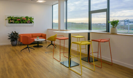 Modern office breakout area with colorful bar stools, a high table, lounge seating, and large windows offering scenic outdoor views