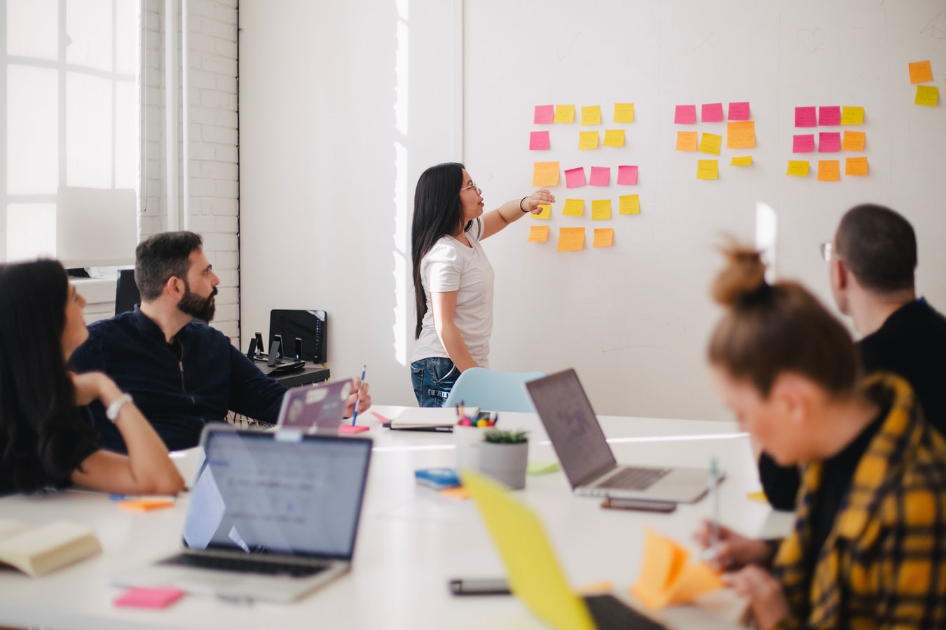 Person pointing at colorful sticky notes on a whiteboard during a team meeting in a bright office, with colleagues seated around a table using laptops and taking notes.