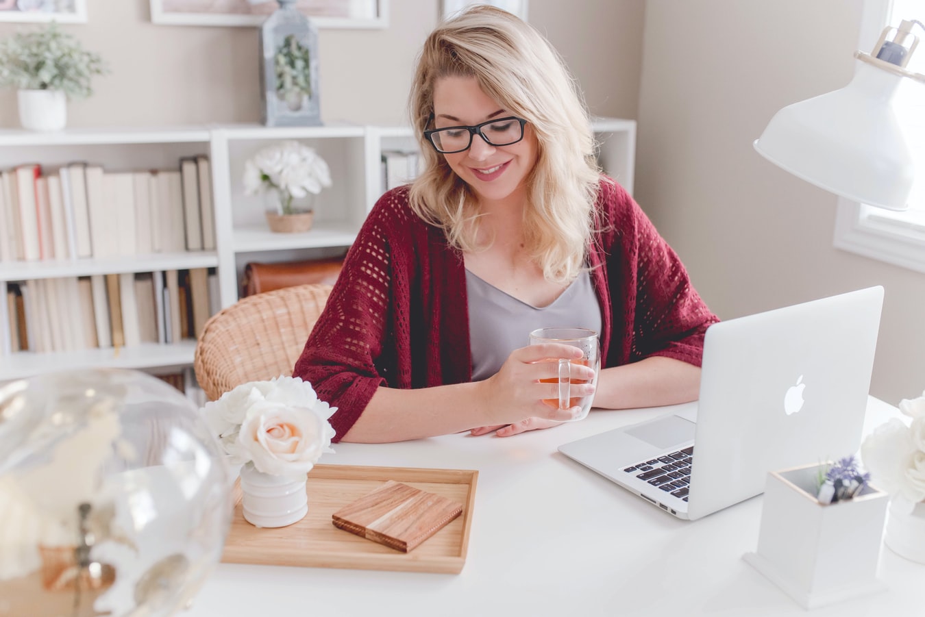 Woman smiling while holding a glass of tea, seated at a desk with a laptop, flowers, and books, in a bright, cozy home office.