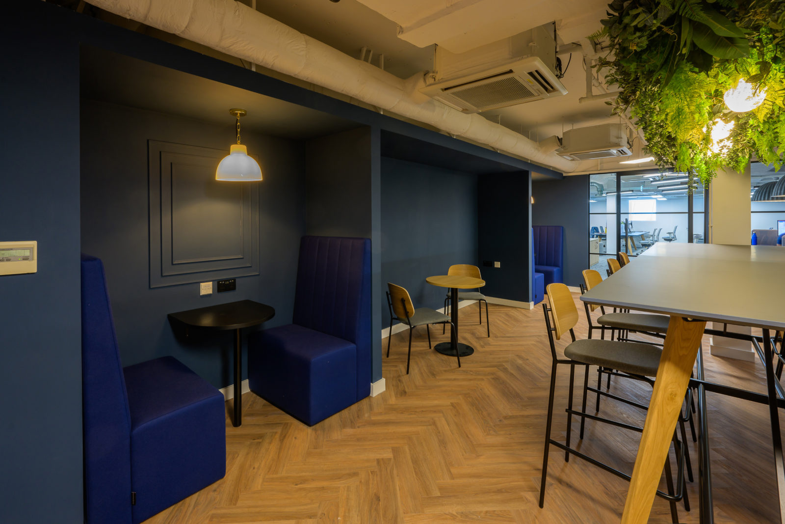 Office lounge with navy blue booth seating and round tables, adjacent to high chairs at a tall table beneath ceiling greenery, illuminated by pendant lights, on herringbone wood flooring.